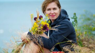 Smiling woman holding wildflowers by the seaside, enjoying summer outdoors.