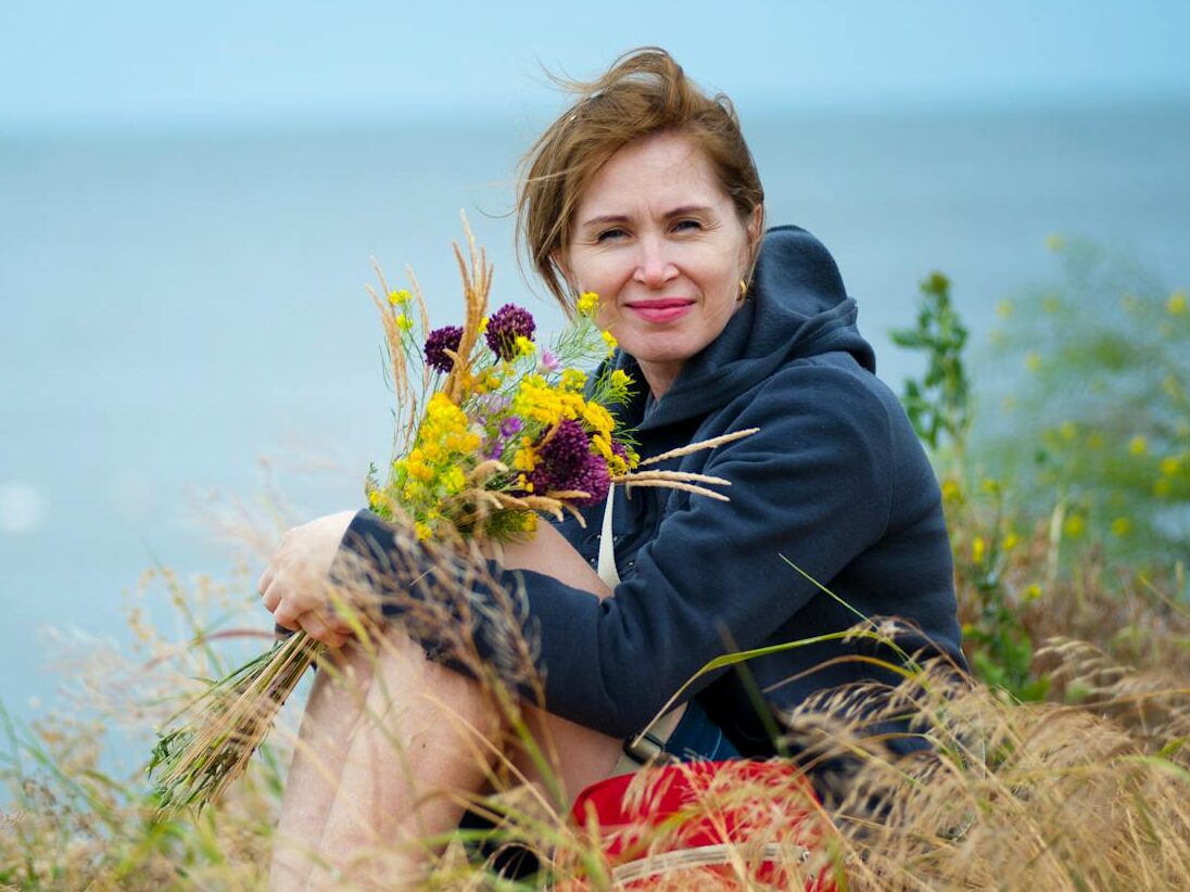 Smiling woman holding wildflowers by the seaside, enjoying summer outdoors.
