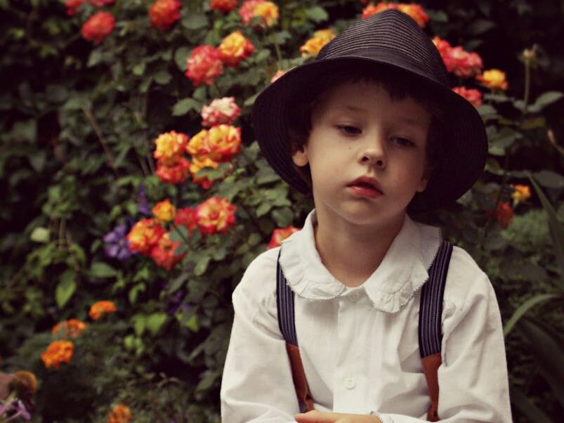 A contemplative boy in vintage attire sits in a garden surrounded by colorful flowers.
