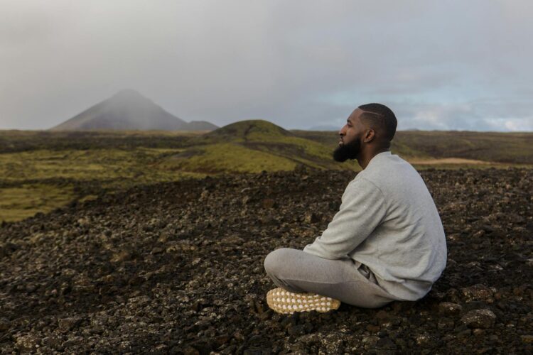 Man meditating on rocky terrain with mountain backdrop. Peace and tranquility outdoors.