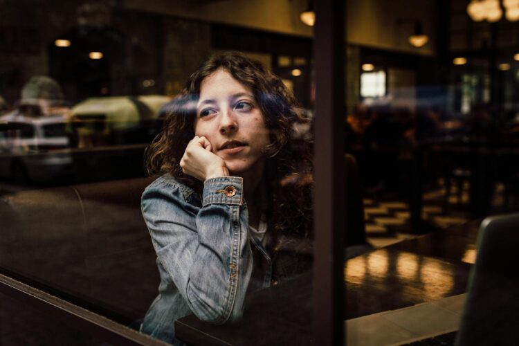 A thoughtful woman sits in a coffee shop, gazing through a window, reflecting in the glass.