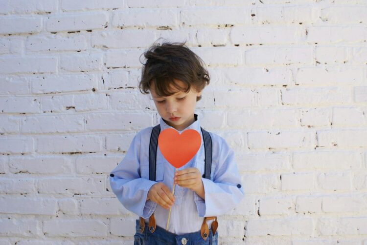 A young boy in suspenders holds a heart-shaped paper against a white brick wall, conveying innocence and emotion.