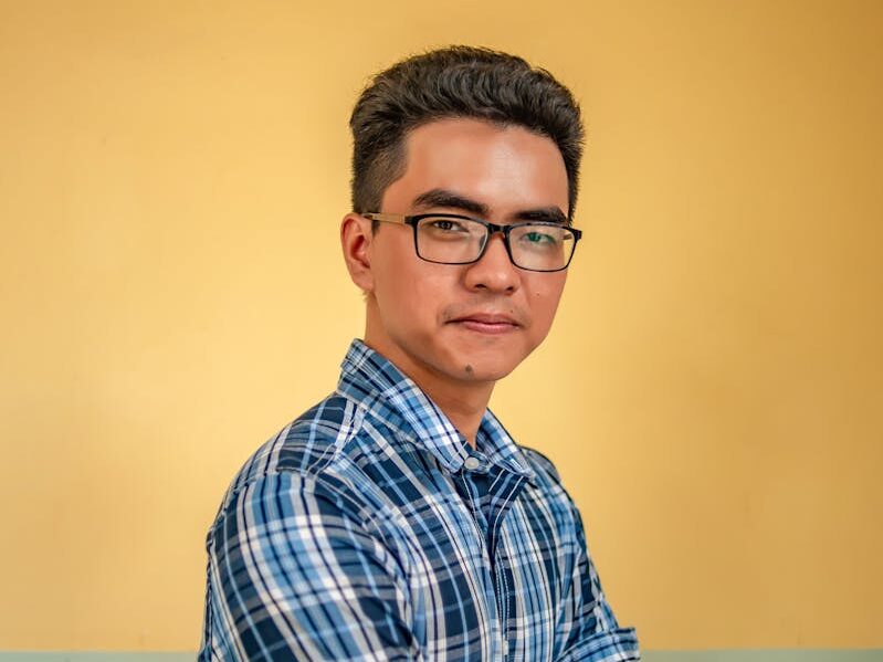A portrait of a confident young man in glasses wearing a plaid shirt against a neutral background.