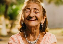Smiling elderly woman with jewelry showcases happiness while holding a mango outdoors.