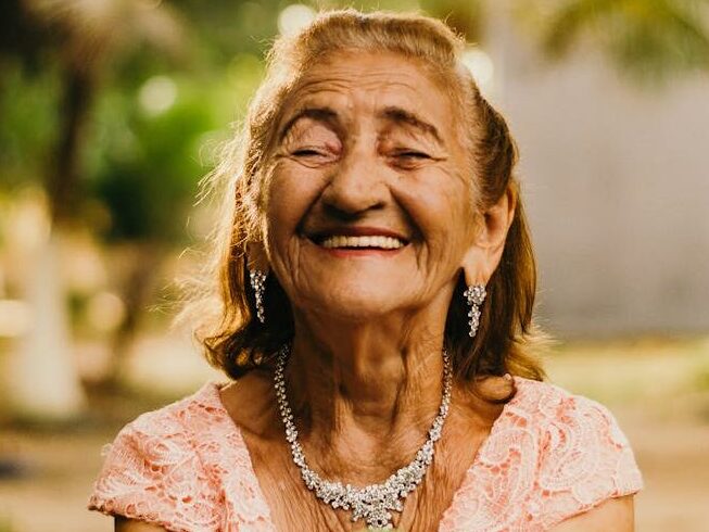 Smiling elderly woman with jewelry showcases happiness while holding a mango outdoors.