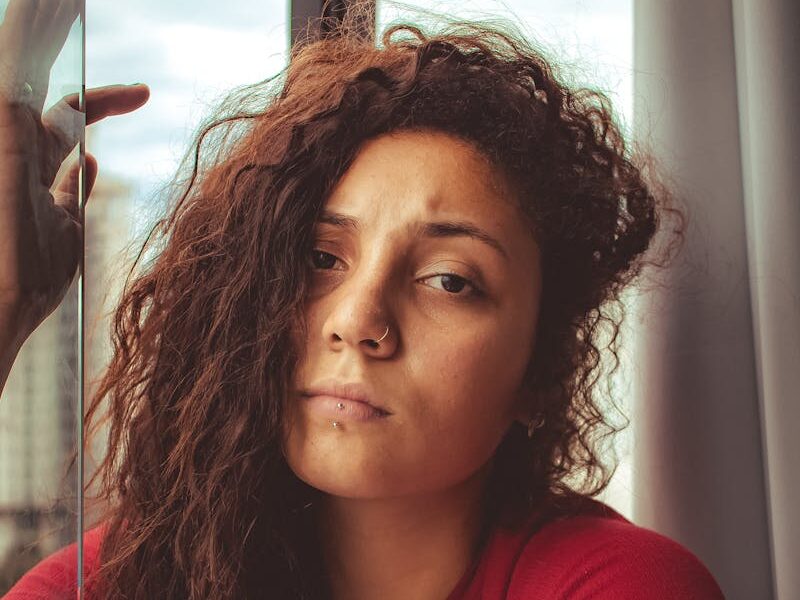 A woman with curly hair looking thoughtfully outside a window in a red striped shirt.