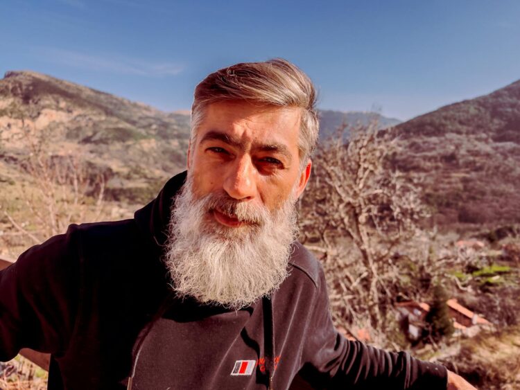 Bearded man poses in scenic Peristera, Greece, with mountains in the backdrop.