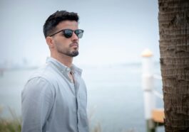 Stylish man in sunglasses gazes thoughtfully by a waterfront, exuding calm and sophistication.