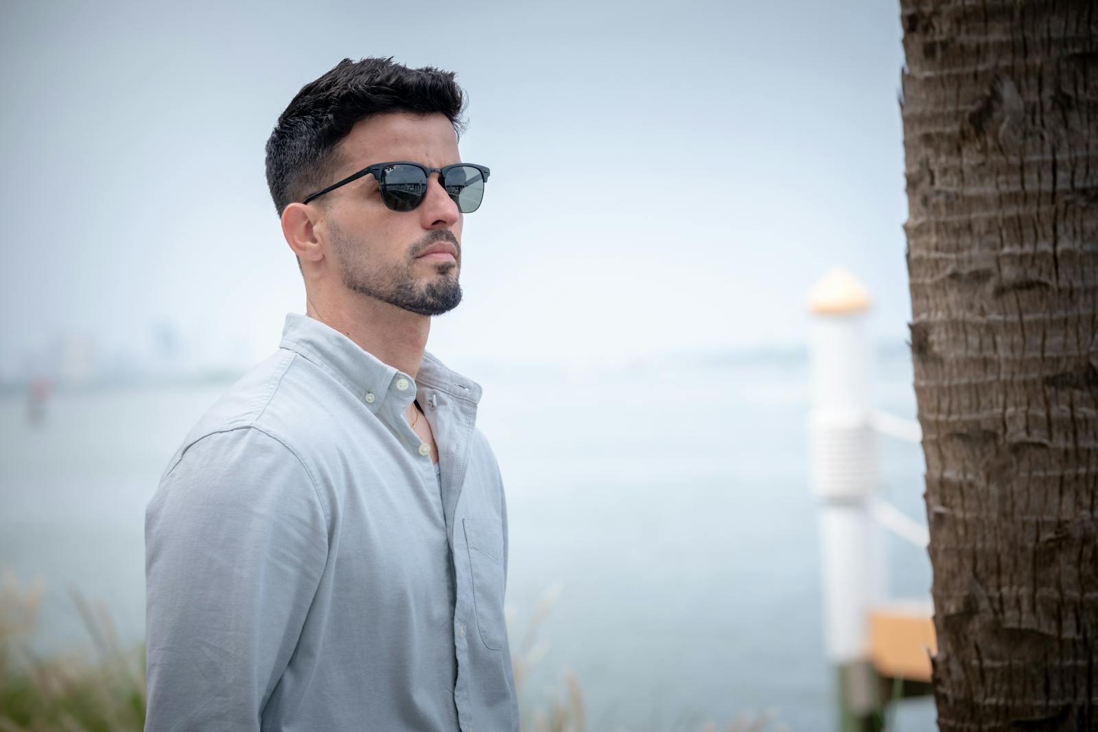 Stylish man in sunglasses gazes thoughtfully by a waterfront, exuding calm and sophistication.