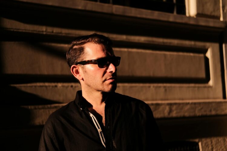 Stylish man wearing sunglasses in dramatic lighting against an architectural backdrop.