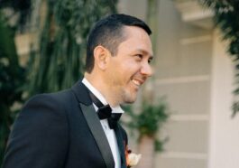 Adult man in a tuxedo smiling outdoors, surrounded by lush greenery, exuding joy.