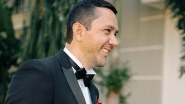 Adult man in a tuxedo smiling outdoors, surrounded by lush greenery, exuding joy.