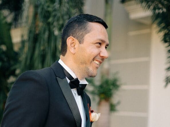 Adult man in a tuxedo smiling outdoors, surrounded by lush greenery, exuding joy.