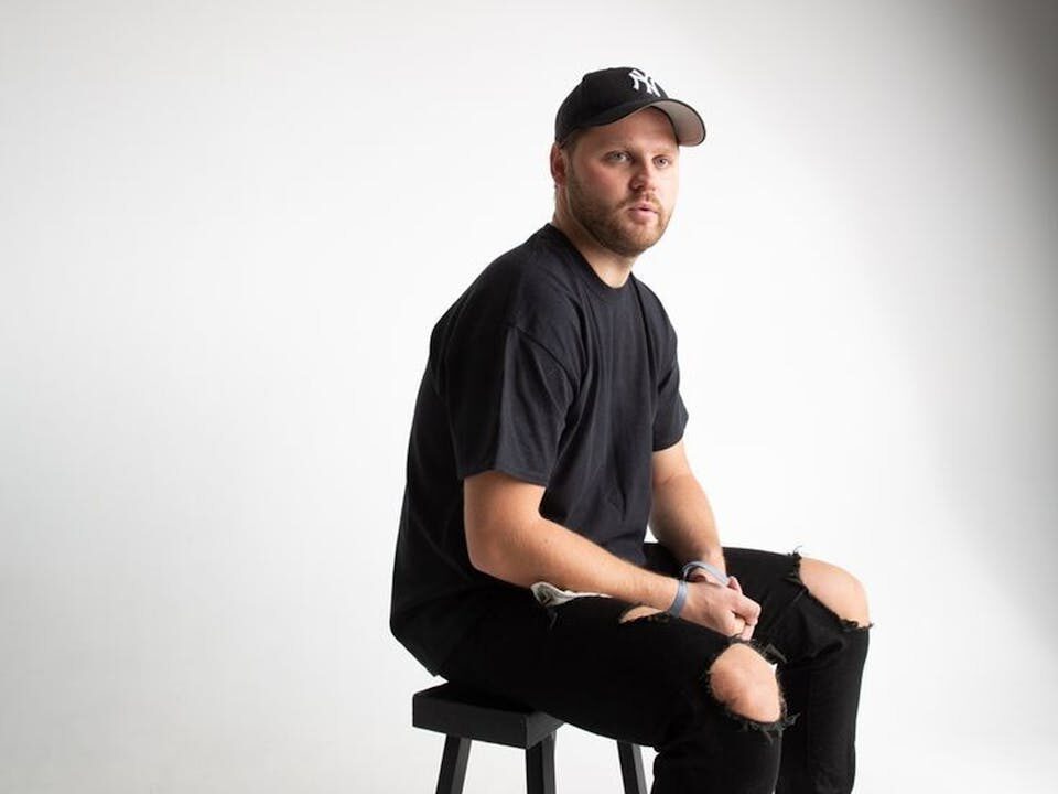 Studio portrait of a seated man wearing casual black outfit and sneakers.