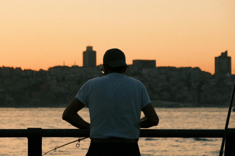 A serene silhouette of a man fishing by the water at sunset, with city skyline in the background.