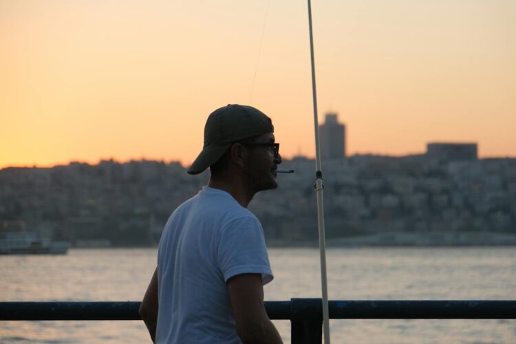 A person fishes at sunset against a city skyline, offering a serene urban coastal scene.