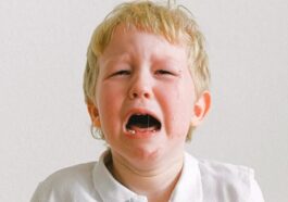 A young child crying, wearing a white shirt, showing strong emotions indoors.