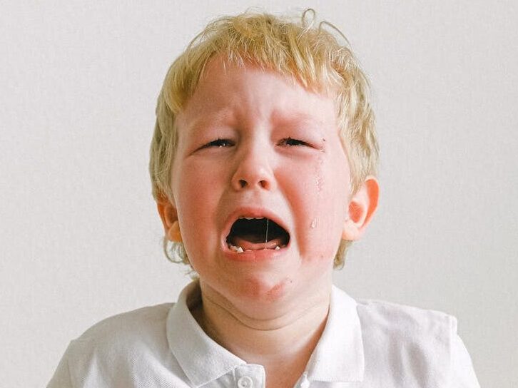 A young child crying, wearing a white shirt, showing strong emotions indoors.