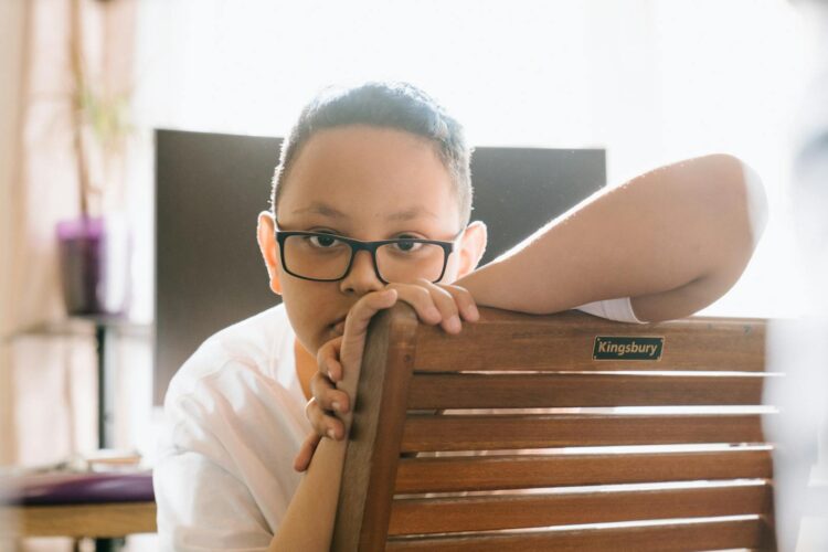 A thoughtful boy wearing glasses sits indoors leaning on a chair.