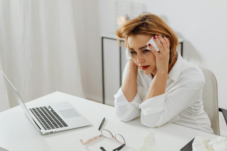 A woman in distress at her office desk, surrounded by tissues and a laptop.