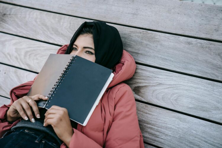 High angle of young Middle East female in casual clothes and traditional headscarf lying on wooden floor of terrace and covering face with notebook
