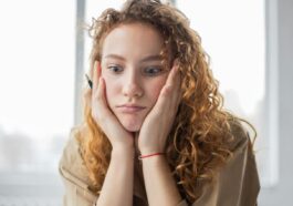 Pensive female student with hands on cheeks looking down while sitting in light room on blurred background while deciding task in frustration