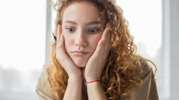 Pensive female student with hands on cheeks looking down while sitting in light room on blurred background while deciding task in frustration
