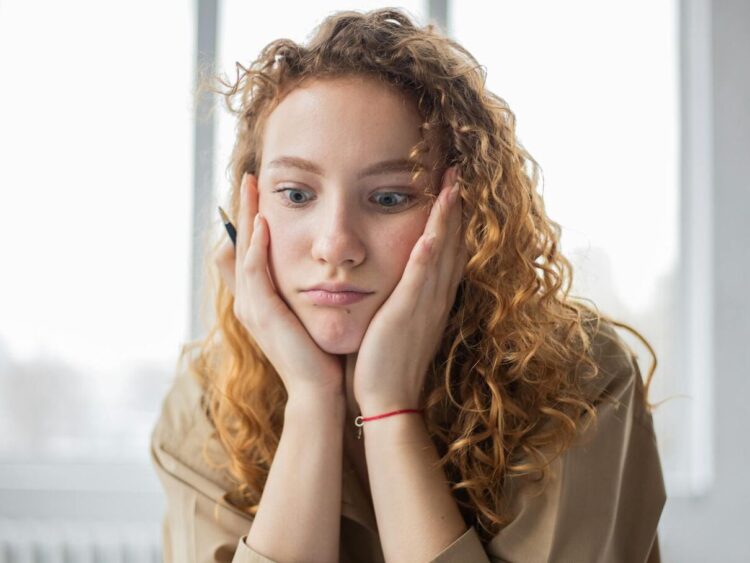 Pensive female student with hands on cheeks looking down while sitting in light room on blurred background while deciding task in frustration