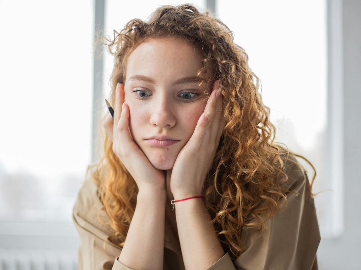 Pensive female student with hands on cheeks looking down while sitting in light room on blurred background while deciding task in frustration
