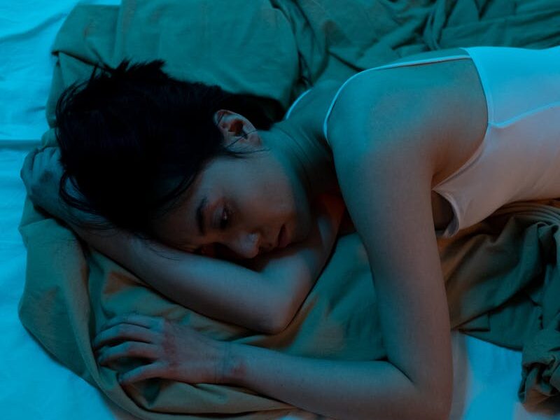 An adult woman lies on a bed in a dimly lit room, reflecting emotions of solitude and contemplation.
