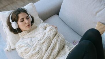 A woman in a cozy sweater relaxes with headphones on a comfortable couch indoors.