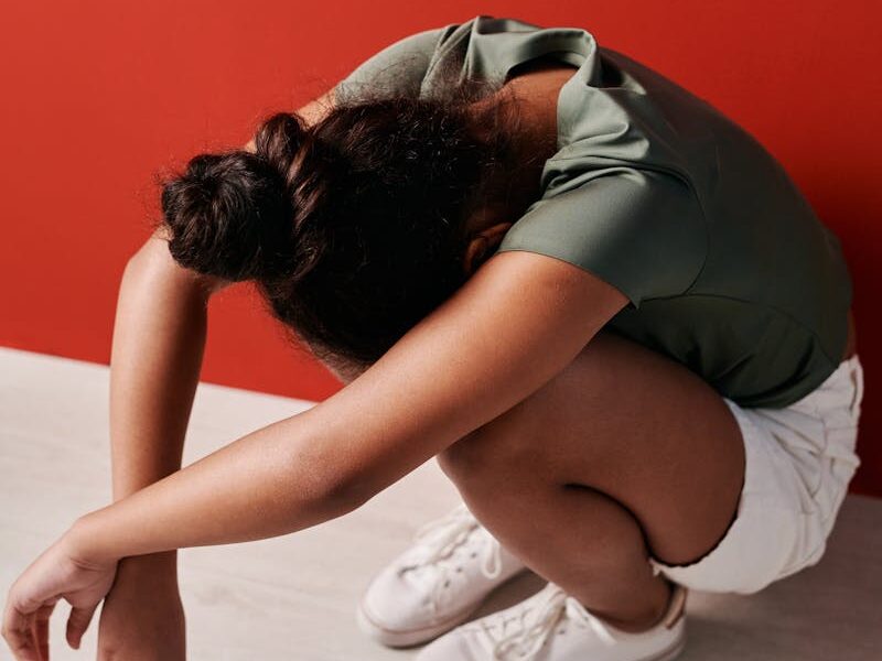 A young girl crouches with her head down wearing white shoes and shorts against a red wall.