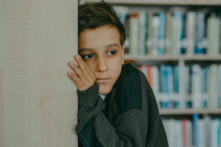 A contemplative child leans against a wall in a library, deep in thought.
