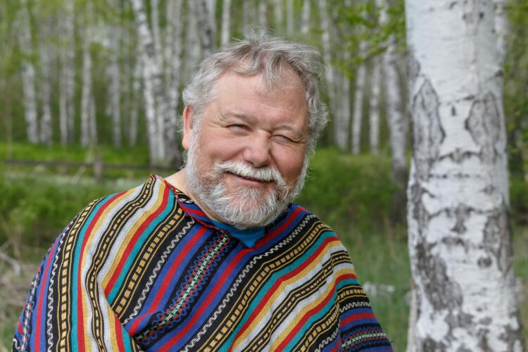 A cheerful senior adult man in a colorful poncho smiling amidst birch trees in a forest setting.