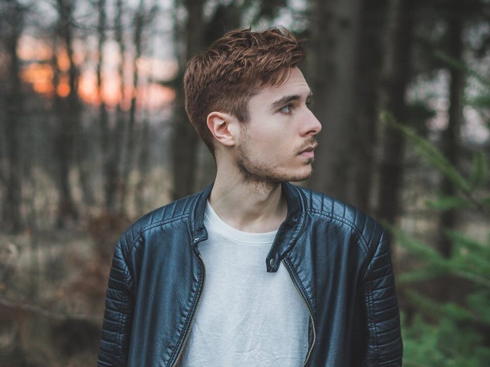 Young man in a black leather jacket standing in a forest during twilight, styled fashion portrait.