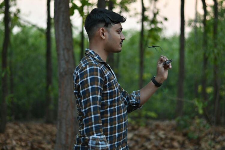 Man in plaid shirt holds glasses in the forest.