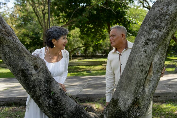 Couple smiles at each other behind two trees.
