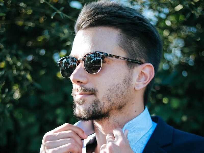 Fashionable man adjusting his suit and sunglasses outdoors on a sunny day.