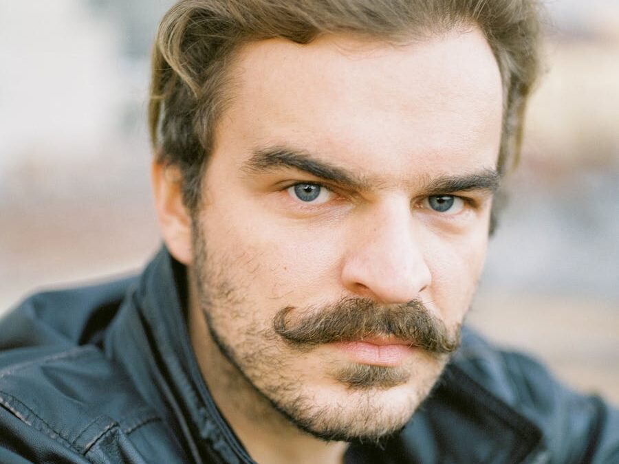 Close-up of a man with a mustache in a leather jacket, looking directly at the camera.