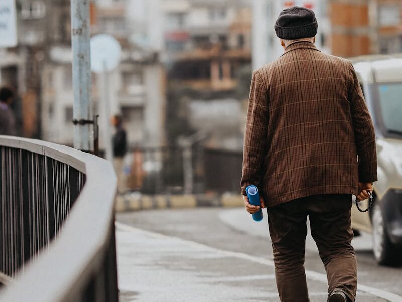 A man walks down a rainy city street holding a blue bottle, clad in a brown jacket and hat.