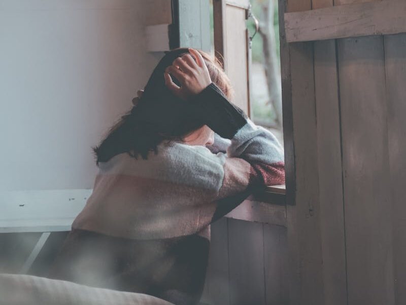 A woman sits indoors gazing thoughtfully out of a window, evoking emotions of loneliness and reflection.