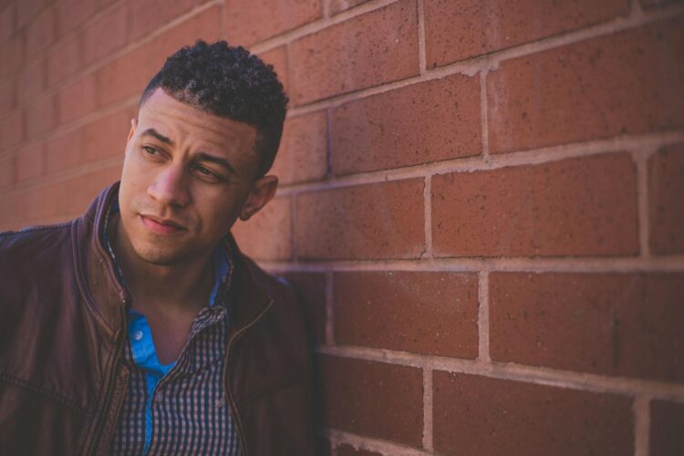 Confident male model in leather jacket posing against a brick wall.