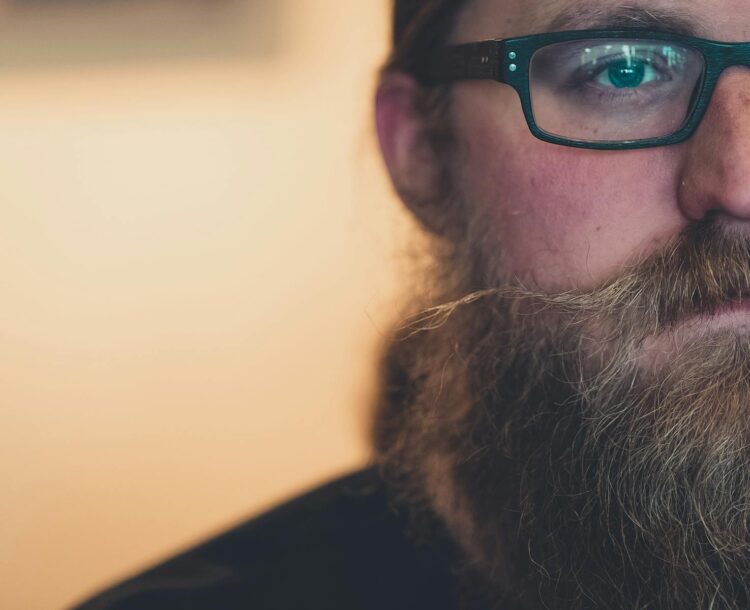 A focused, artistic close-up of a bearded man wearing eyeglasses, emphasizing facial features.