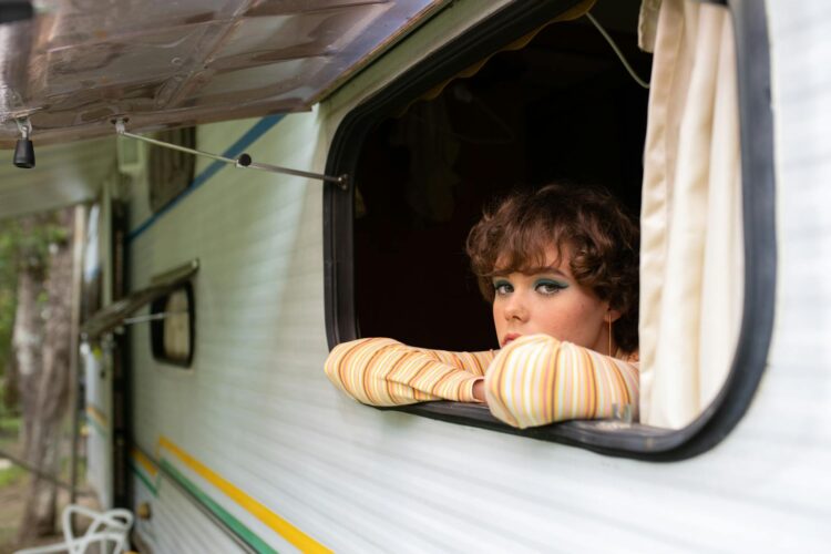 A young woman with makeup relaxes in a campervan, gazing outside on a summer day.