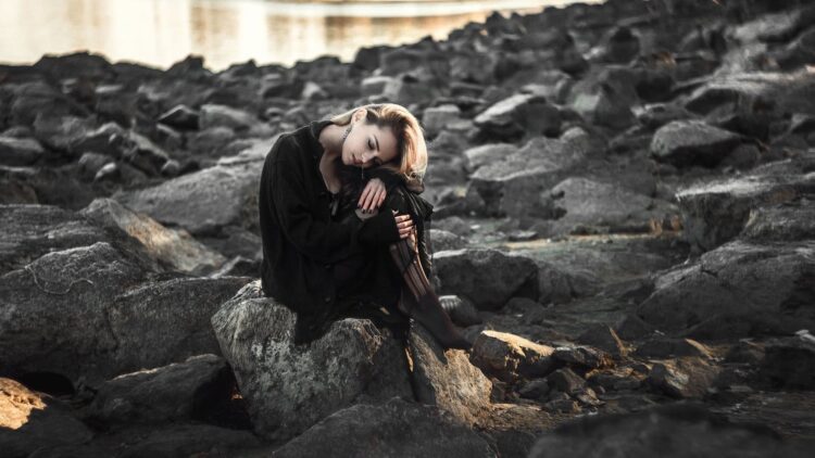 A woman in a black outfit sitting on rocks by the water, reflective mood.
