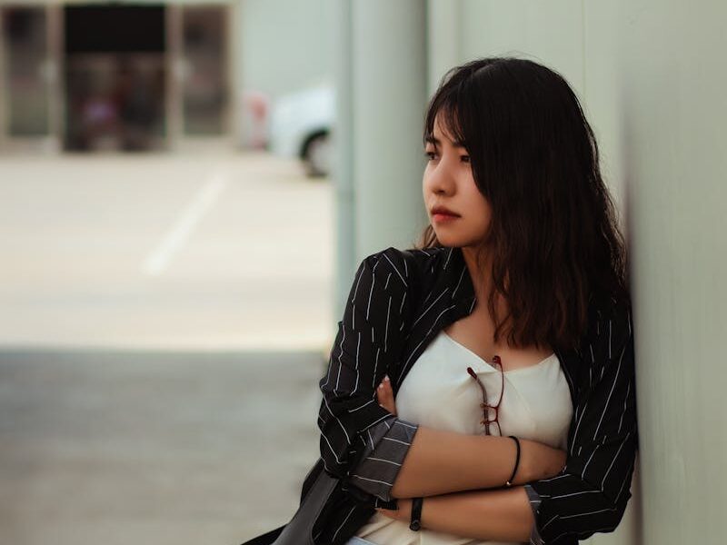 A young woman leans against a wall outdoors, deep in thought, in a casual urban setting.