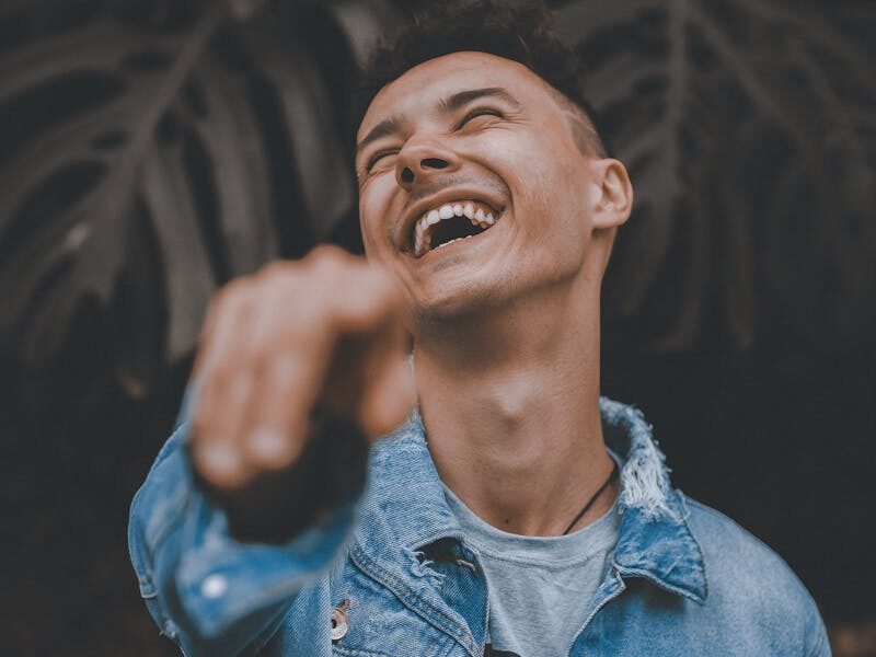 A joyful man in a denim jacket laughs and points, surrounded by lush greenery.
