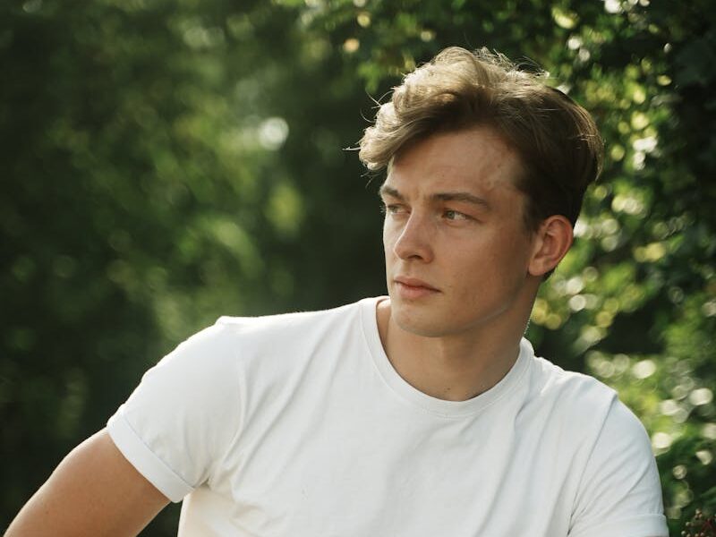 Portrait of a young man sitting outdoors in white t-shirt and plaid pants, enjoying a sunny day.