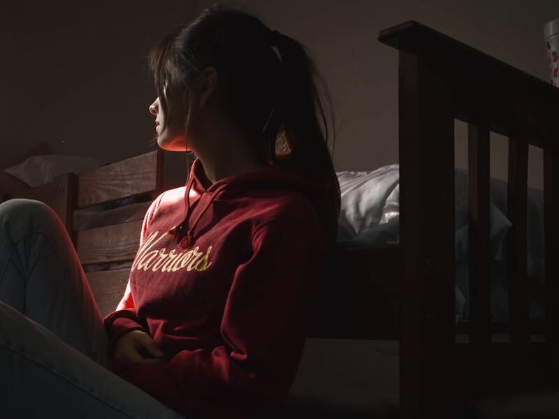 Contemplative young woman in hoodie sitting on the floor in dimly lit room, symbolizing solitude and introspection.