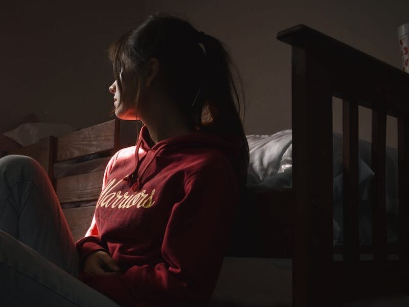 Contemplative young woman in hoodie sitting on the floor in dimly lit room, symbolizing solitude and introspection.